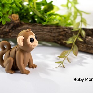 May include: A small brown toy monkey with large black eyes and a curled tail. The monkey is sitting on a white surface with a branch and green leaves in the background. The text "Baby Monkey" is written below the monkey.