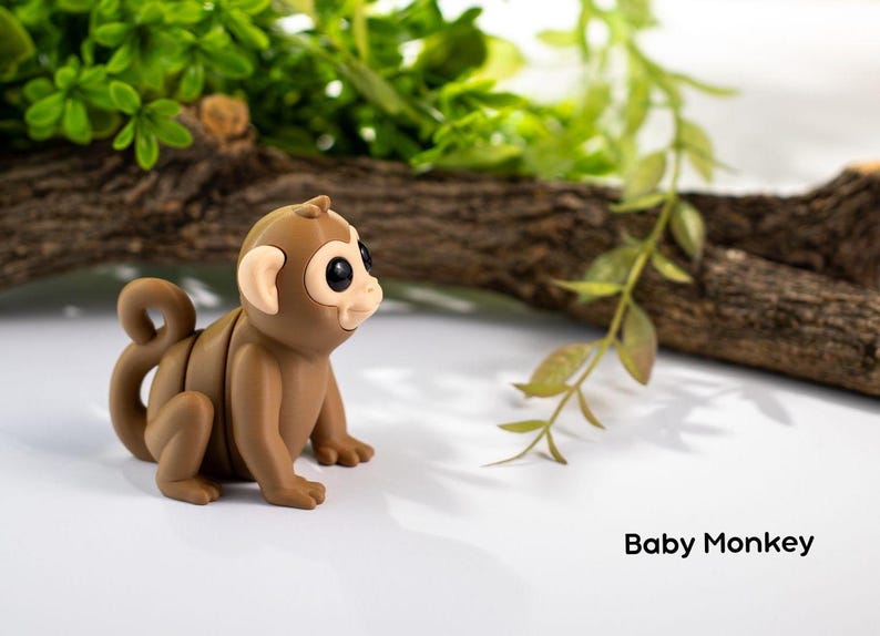 May include: A small brown toy monkey with large black eyes and a curled tail. The monkey is sitting on a white surface with a branch and green leaves in the background. The text "Baby Monkey" is written below the monkey.