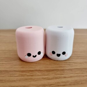 Op de afbeelding: Twee kleine, ronde, keramische beeldjes in de vorm van marshmallows met schattige gezichtjes. De marshmallow aan de linkerkant is roze en de marshmallow aan de rechterkant is wit. Beide marshmallows hebben zwarte ogen, een zwarte glimlach en roze wangen.
