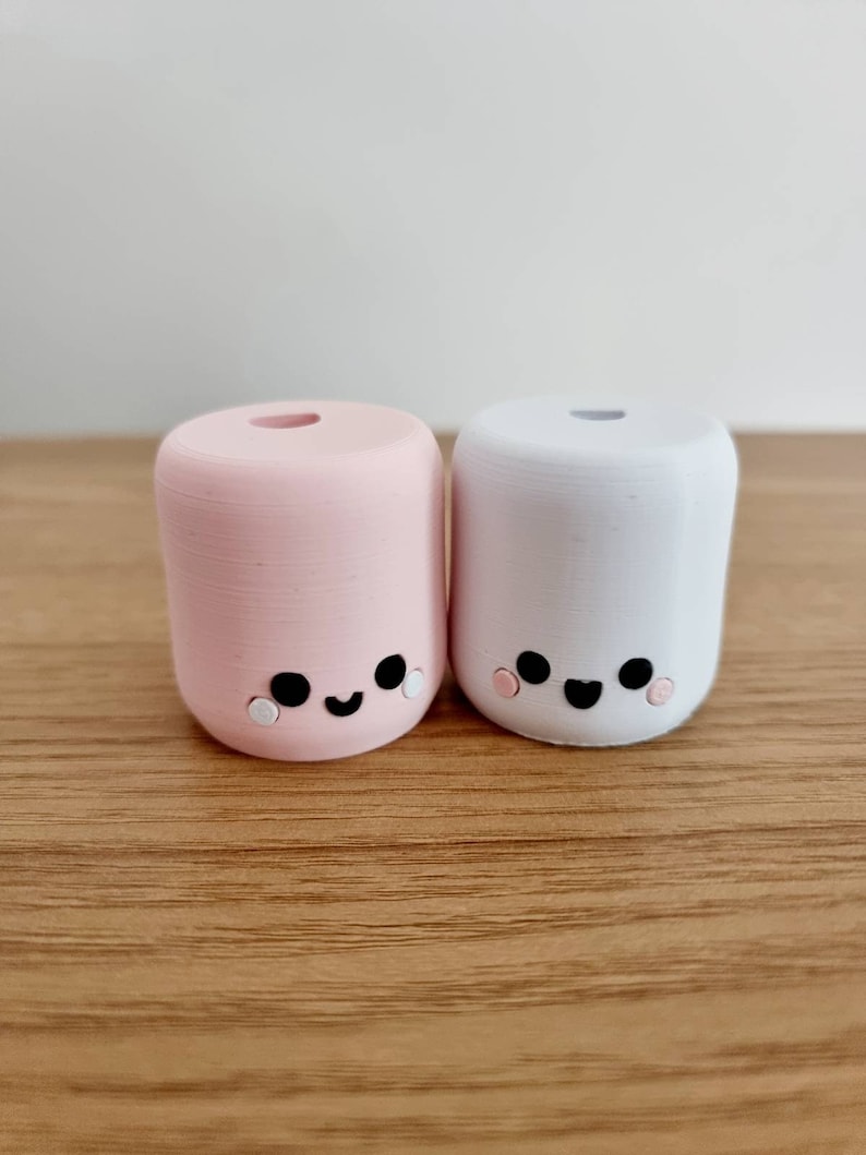 Op de afbeelding: Twee kleine, ronde, keramische beeldjes in de vorm van marshmallows met schattige gezichtjes. De marshmallow aan de linkerkant is roze en de marshmallow aan de rechterkant is wit. Beide marshmallows hebben zwarte ogen, een zwarte glimlach en roze wangen.