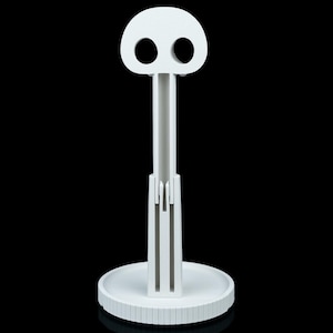 May include: A white, stylized paper towel holder with a skull-like top. The holder has a circular base with a ridged edge, a central support, and a top with two eye-shaped holes. The design is minimalist and modern.