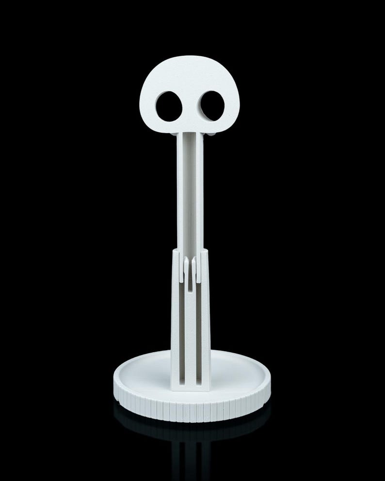 May include: A white, stylized paper towel holder with a skull-like top. The holder has a circular base with a ridged edge, a central support, and a top with two eye-shaped holes. The design is minimalist and modern.