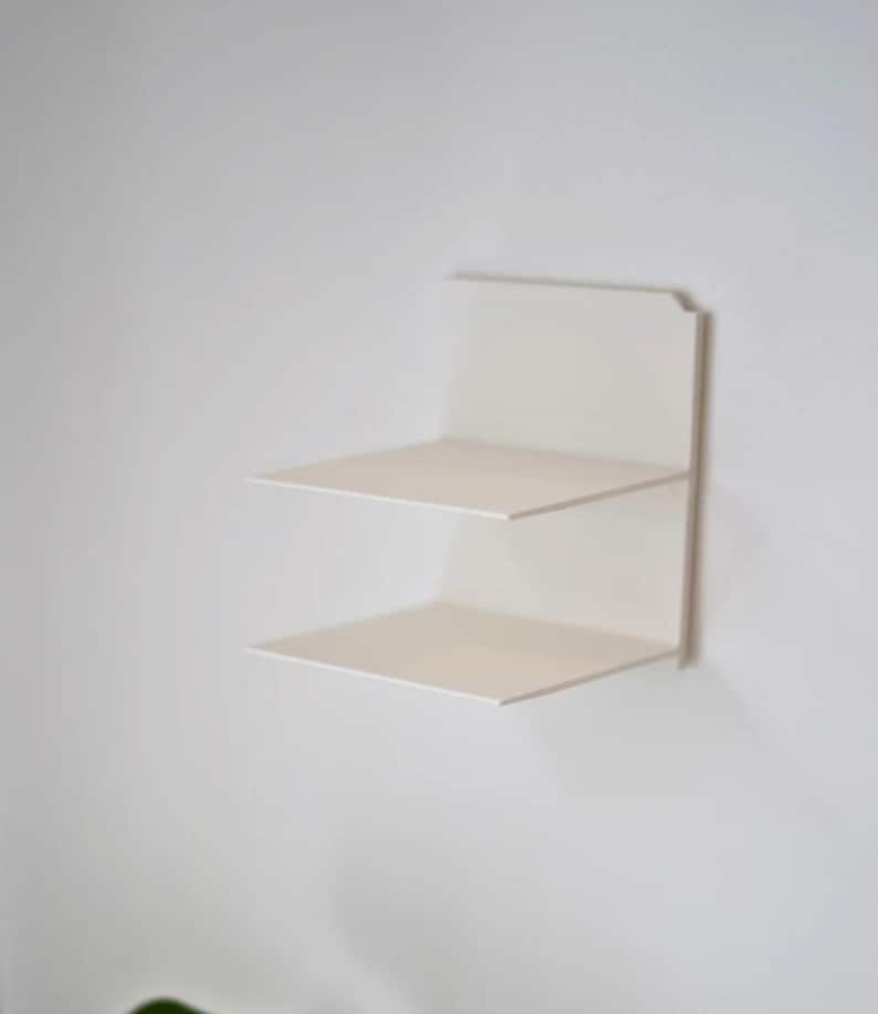 May include: A minimalist, white, two-tiered wall shelf. The shelf has a clean, modern design with two rectangular platforms. The shelf is mounted on a white wall, creating a simple and functional storage solution.