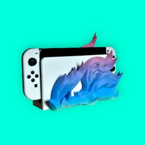 May include: A white Nintendo Switch console with a blue and pink 3D printed stand shaped like a flame. The stand is designed to hold the console upright.