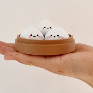 May include: Three white steamed buns with cute faces are arranged in a brown bamboo steamer. The steamer is sitting on a person's hand.