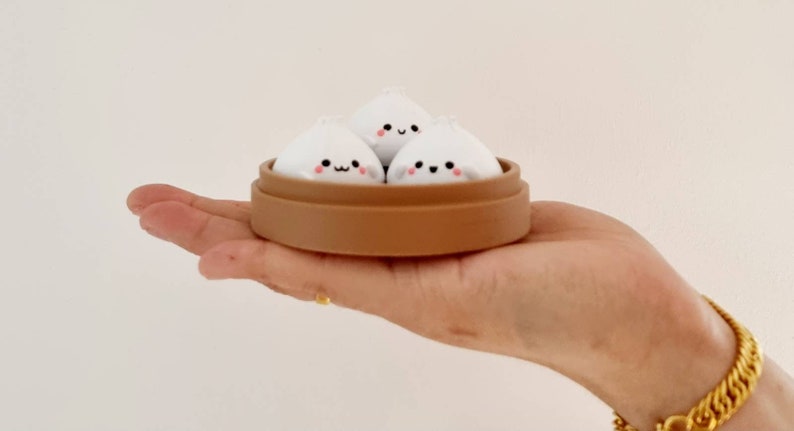 May include: Three white steamed buns with cute faces are arranged in a brown bamboo steamer. The steamer is sitting on a person's hand.