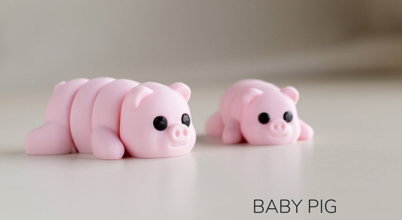 Puede incluir: Dos peque&ntilde;as figuras de cerdos rosas con ojos negros y caras sonrientes. Un cerdo est&aacute; segmentado, el otro es de una sola pieza. El texto "BABY PIG" est&aacute; debajo.