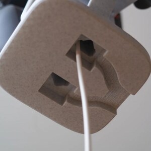 May include: Close-up of a light beige, textured, square-shaped object with a rectangular cutout and a white cord extending from it. The object appears to be part of a larger structure, possibly a lamp or electronic device.