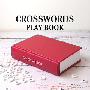 Puede incluir: Un libro de juegos de crucigramas rojo con la palabra "CROSSWORDS" impresa en la portada. Las fichas de letras blancas est&aacute;n esparcidas alrededor del libro. El texto "CROSSWORDS PLAY BOOK" est&aacute; encima del libro.