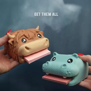 以下が含まれることがあります： 茶色のハイランド牛とターコイズ色のカバの、2つの動物型付箋ホルダー。それぞれピンク色の付箋パッドが付いています。「GET THEM ALL」の文字が牛の上にあります。