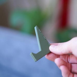 Może przedstawiać: Mały, oliwkowozielony stojak na telefon. Stojak wykonany jest z solidnego materiału i ma tr&oacute;jkątny kształt z p&oacute;łką do trzymania telefonu. Stojak jest trzymany w dłoni.