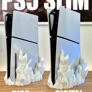 May include: Two white PS5 Slim consoles, one with a disc drive and one digital, are displayed on a wooden surface. Both consoles are on a white crystal-shaped stand.