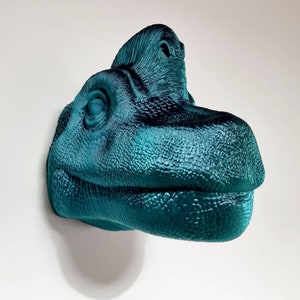 May include: Teal blue dinosaur head wall sculpture with a textured surface. The dinosaur has a large, open mouth and a small, pointed nose.