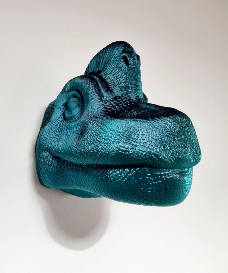 May include: Teal blue dinosaur head wall sculpture with a textured surface. The dinosaur has a large, open mouth and a small, pointed nose.