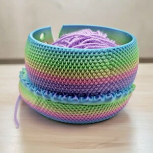 May include: A rainbow yarn bowl with a purple yarn ball inside. The bowl is made of plastic and has a textured surface.