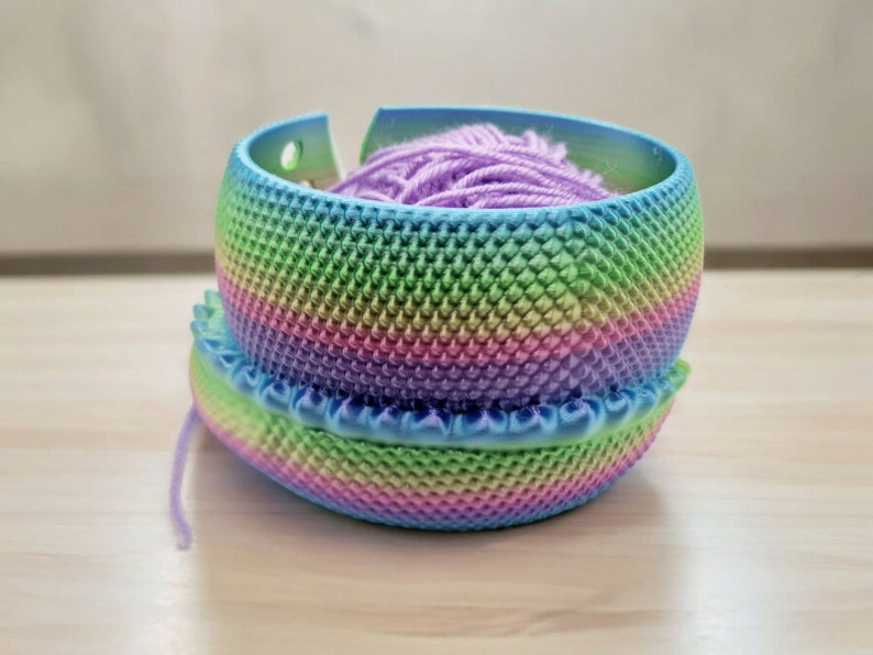 May include: A rainbow yarn bowl with a purple yarn ball inside. The bowl is made of plastic and has a textured surface.