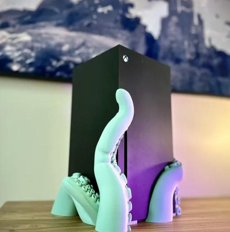 May include: A teal blue 3D printed octopus stand holds a black gaming console. The console has a white button on the top.