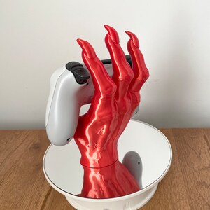 May include: A red, 3D printed hand holding a white video game controller. The hand is posed with claws and is on a rotating base.