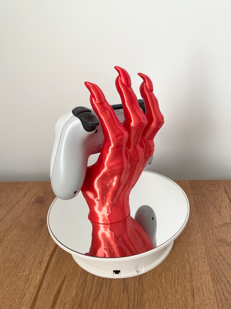 May include: A red, 3D printed hand holding a white video game controller. The hand is posed with claws and is on a rotating base.