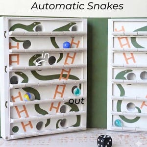 May include: A white wooden game with green snake illustrations, orange ladders, and the text "Automatic Snakes." The game includes colorful marbles and a black and white die. The word "in" and "out" are also visible.