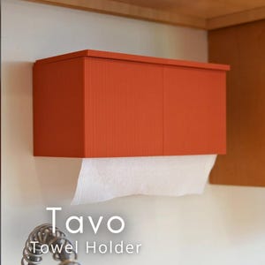 May include: An orange paper towel holder mounted on a white wall. The holder has a ridged texture and the text "Tavo Towel Holder" is displayed below. White paper towels are dispensed from the bottom.