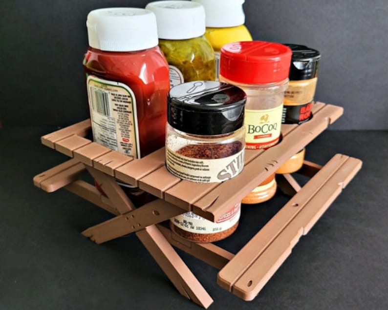 May include: A brown wooden picnic table-style spice rack with six glass jars filled with various spices. The jars have red, black, and white lids. The spices include ketchup, mustard, and various seasonings.