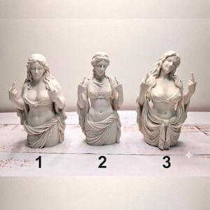 May include: Three off-white statues of female figures in classical drapery, each displaying a middle finger gesture. The figures are numbered 1, 2, and 3 at the base. Each bust features detailed hair and fabric folds, presented in a uniform matte finish.