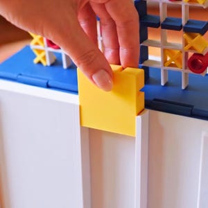 May include: A plastic board game featuring a white grid structure with blue accents. The game includes yellow square tiles and red cylindrical pieces. The board has a white base and a vertical grid section where players insert the colored game pieces.
