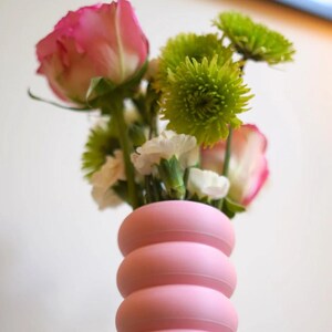 May include: A pink vase with a stacked, rounded design holds a bouquet of flowers. The bouquet includes pink roses, green chrysanthemums, and white carnations. The vase is a soft, pastel pink color.