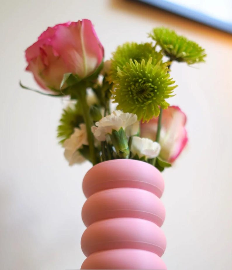 May include: A pink vase with a stacked, rounded design holds a bouquet of flowers. The bouquet includes pink roses, green chrysanthemums, and white carnations. The vase is a soft, pastel pink color.