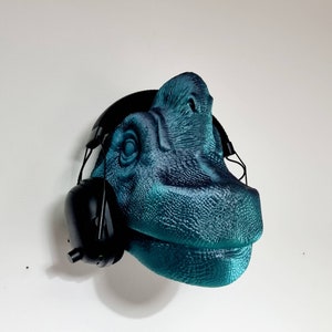 May include: A teal blue dinosaur head sculpture wearing black headphones. The dinosaur has a textured surface and a wide open mouth.