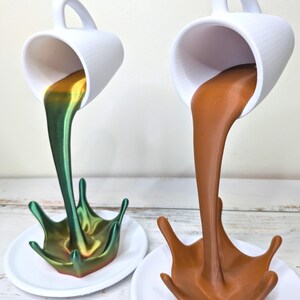May include: Two white ceramic mugs appear to be pouring liquid into a splash shape. One mug pours a green and gold liquid, the other a brown liquid. Both are on white saucers, creating a unique decorative display.