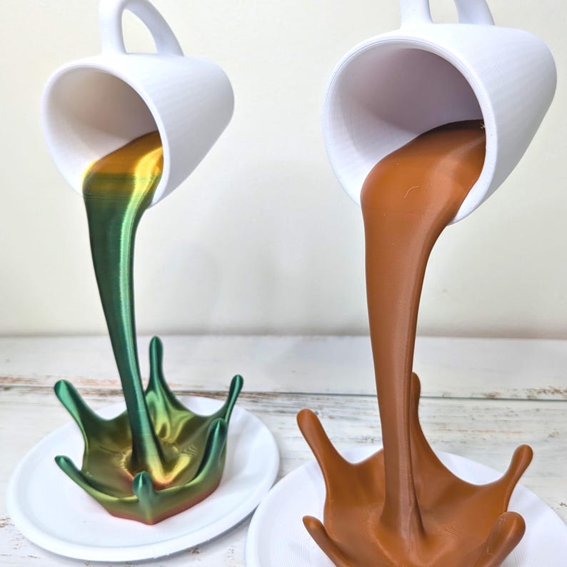 May include: Two white ceramic mugs appear to be pouring liquid into a splash shape. One mug pours a green and gold liquid, the other a brown liquid. Both are on white saucers, creating a unique decorative display.