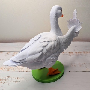 May include: A white goose ornament with outstretched wings, an orange beak and feet, and a green base. The goose has detailed feather textures. The ornament is a decorative item.