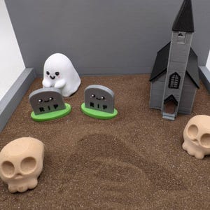Pu&ograve; includere: Una scena in miniatura a tema Halloween con un fantasma bianco, due lapidi grigie con la scritta "RIP", una chiesa grigia e un teschio color sabbia su una superficie di sabbia marrone.