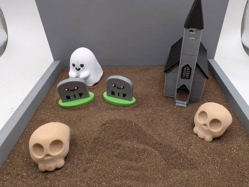 Pu&ograve; includere: Una scena in miniatura a tema Halloween con un fantasma bianco, due lapidi grigie con la scritta "RIP", una chiesa grigia e un teschio color sabbia su una superficie di sabbia marrone.