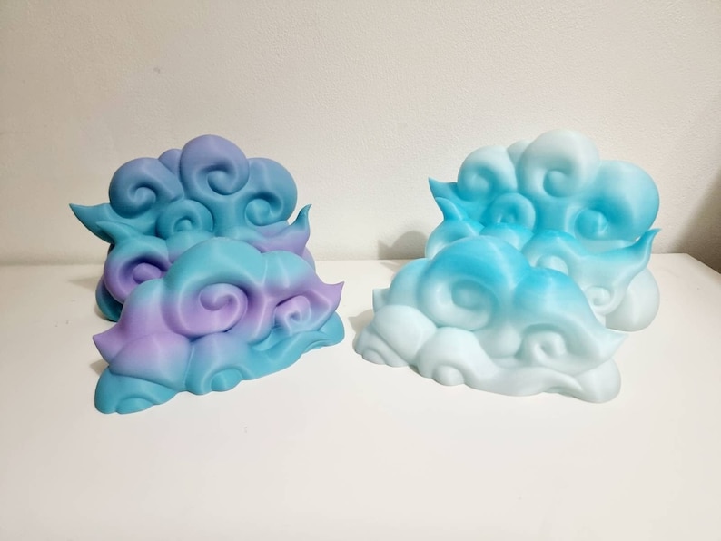 May include: Four decorative cloud-shaped objects in shades of blue, purple, and white. The clouds have a swirling design and are arranged on a white surface. The objects appear to be 3D-printed.