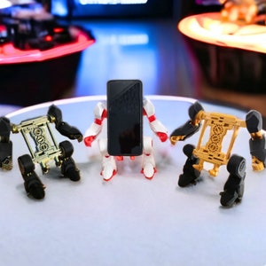 May include: Three robot figures holding a black smartphone. The robots are made of plastic and have a white, red, and gold color scheme. The robots are standing on a white surface.