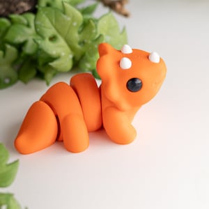 May include: Orange and white 3D printed toy dinosaur with black eyes. The dinosaur is curled up in a playful pose.