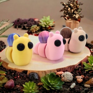 May include: Three colorful, hand-painted, ceramic snail figurines. The snails are pink, yellow, and white with black eyes. The snails are sitting on a wooden log with green plants around them.