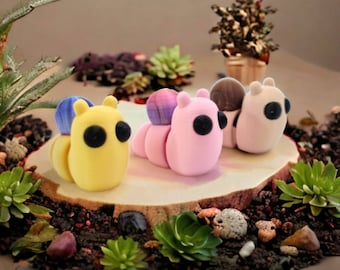 May include: Three colorful, hand-painted, ceramic snail figurines. The snails are pink, yellow, and white with black eyes. The snails are sitting on a wooden log with green plants around them.