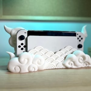 Könnte beinhalten: Ein weißer Nintendo Switch Konsolenständer mit Wolken-Design. Der Ständer ist aus Kunststoff und hat einen Ausschnitt für den Bildschirm der Konsole. Der Ständer ist so konzipiert, dass er die Konsole aufrecht hält und einen Platz für die Joy-Con-Controller der Konsole bietet.