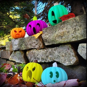 May include: Five colorful, carved pumpkin decorations with different facial expressions. The pumpkins are arranged on a stone wall with autumn leaves scattered around them.