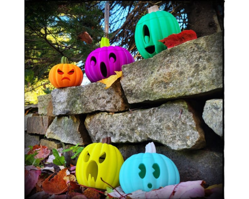 May include: Five colorful, carved pumpkin decorations with different facial expressions. The pumpkins are arranged on a stone wall with autumn leaves scattered around them.