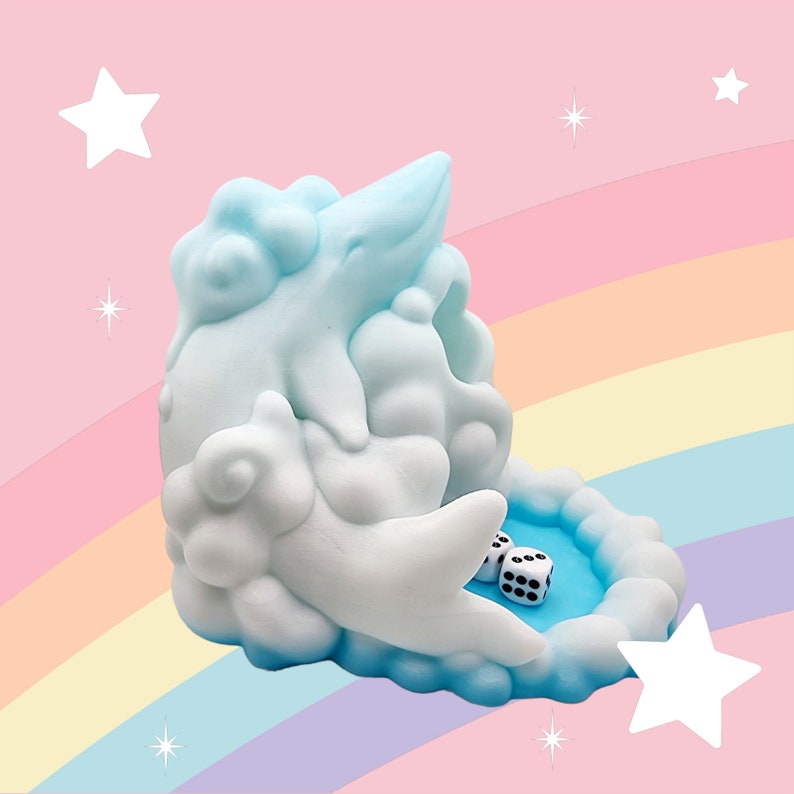 Puede incluir: Una bandeja de dados caprichosa en forma de nube, en tonos azules y blancos, con un interior azul claro. Dos dados blancos con puntos negros descansan dentro de la bandeja. El fondo presenta un arco&iacute;ris y estrellas.