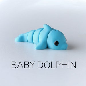 May include: A small, light blue plastic toy dolphin with black eyes. The dolphin is lying on its side with its head turned to the right. The text "BABY DOLPHIN" is printed below the dolphin.