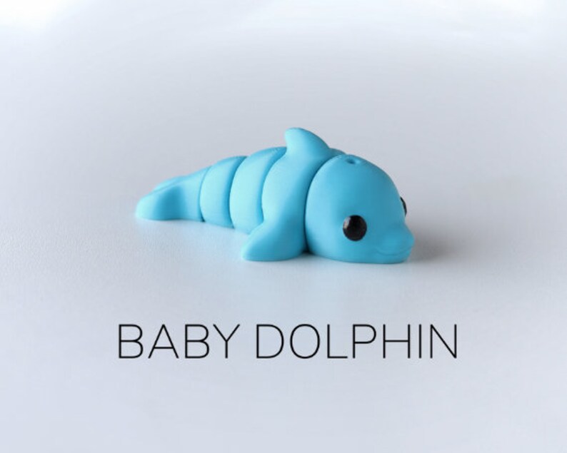 May include: A small, light blue plastic toy dolphin with black eyes. The dolphin is lying on its side with its head turned to the right. The text "BABY DOLPHIN" is printed below the dolphin.