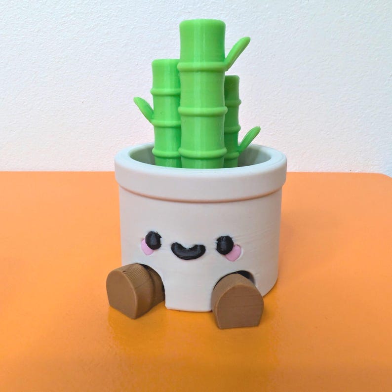 May include: A whimsical, off-white planter with a smiling face, pink cheeks, and brown feet, holding bright green bamboo stalks. The planter sits on an orange surface, creating a cheerful, decorative accent.