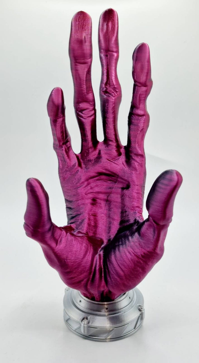May include: A 3D printed hand sculpture with a metallic silver base. The hand is made of a deep pink, almost magenta, plastic and is posed with the palm facing forward.