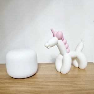 May include: A white ceramic unicorn figurine with pink balloon-like details. The unicorn is standing on a wooden surface next to a white round smart home device.